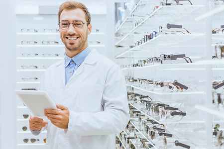 Professional smiling optometrist using digital tablet near eyeglasses on shelves in ophthalmic shopの写真素材