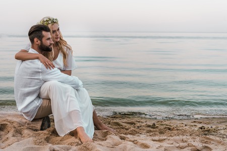 Elegant groom and bride sitting on log on beach and looking awayの写真素材