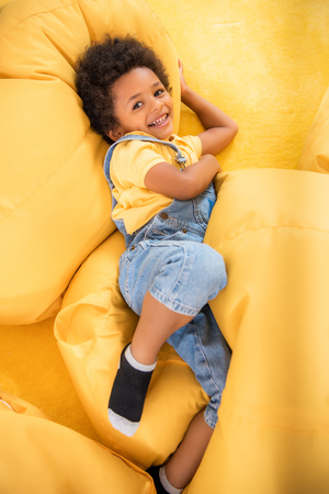 High angle view of smiling African american boy lying on bean bag chairs and looking at camera in kindergartenの写真素材