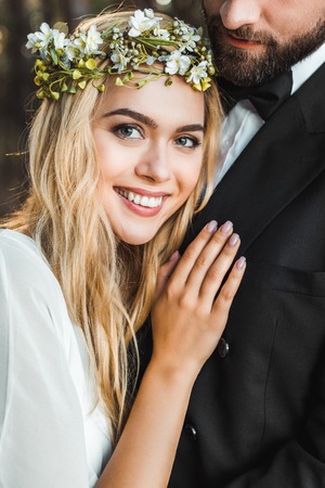 Smiling attractive bride in wreath hugging groom and looking at cameraの写真素材