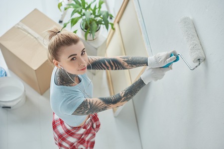 High angle view of young tattooed woman in earphones painting wall at new homeの写真素材