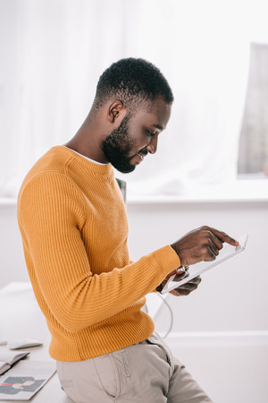 Side view of handsome African American designer in orange sweater using tablet in officeの写真素材