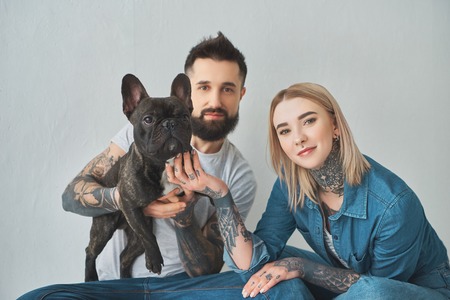 Young tattooed couple holding french bulldog and smiling at camera on greyの写真素材
