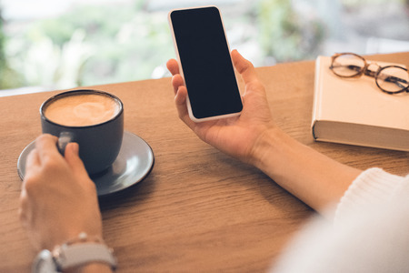Cropped image of woman with book and coffee cup using smartphone at table in cafe