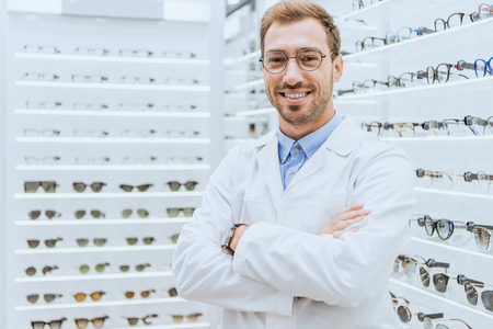 Portrait of smiling professional optometrist in glasses posing with crossed arms in ophthalmic shop