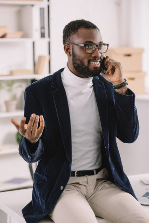 Bearded stylish African American businessman talking smartphone on while sitting on table in officeの写真素材