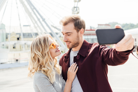 Smiling couple in autumn outfit looking at each other and taking selfie with smartphone in cityの写真素材
