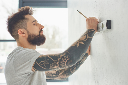 Side view of bearded young man holding level tool and marking wall with pencil during repairsの写真素材