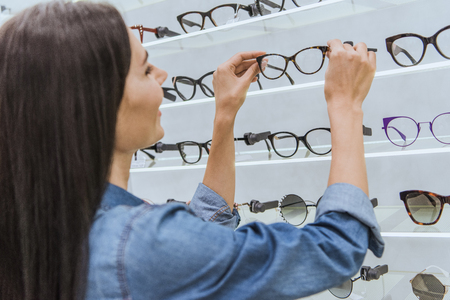 Side view of attractive young woman taking eyeglasses from shelves in ophthalmic shopの写真素材