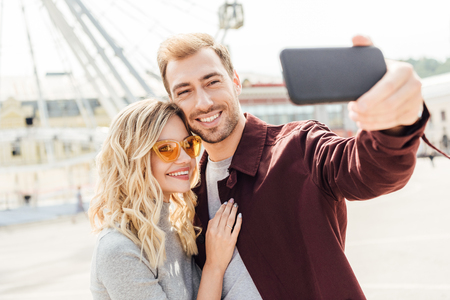 Smiling couple in autumn outfit taking selfie with smartphone in cityの写真素材