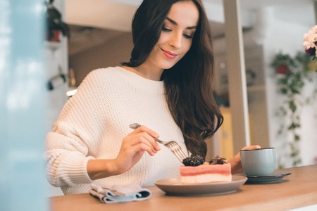 Selective focus of young woman with coffee cup eating cheesecake at table in cafeの写真素材