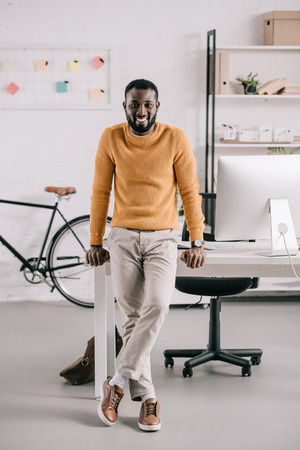 Smiling handsome African American designer in orange sweater leaning on table and looking at camera in officeの写真素材