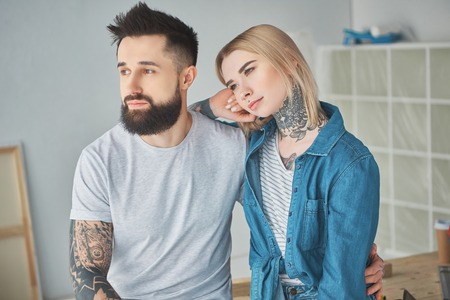 Beautiful young couple with tattoos sitting together and looking away in new houseの写真素材