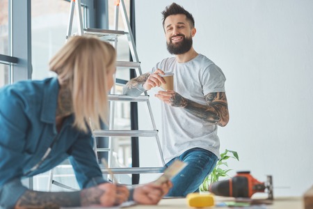 Happy bearded man holding paper cup and looking at girlfriend on foreground in new houseの写真素材