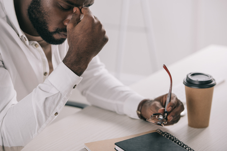 Tired African American businessman touching nose bridge and holding glasses in officeの写真素材