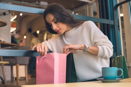 Focused young woman looking in shopping bag at table in cafeの写真素材