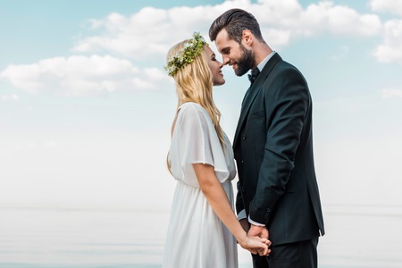 Wedding couple in suit and white dress holding hands and touching with noses on beachの写真素材