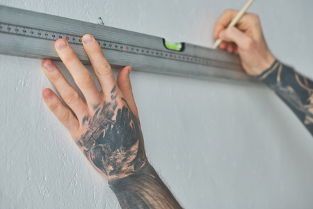 Cropped shot of tattooed man holding level tool and marking wall with pencil during repairs