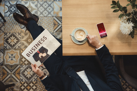 Overhead view of businessman with newspaper sitting at table with cup of coffee and smartphone in cafeの写真素材