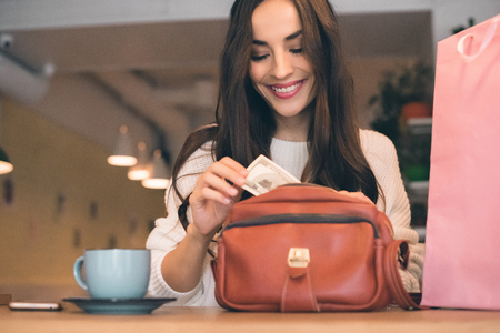 Smiling woman pulling out cash from bag at table with coffee cup in cafeの写真素材