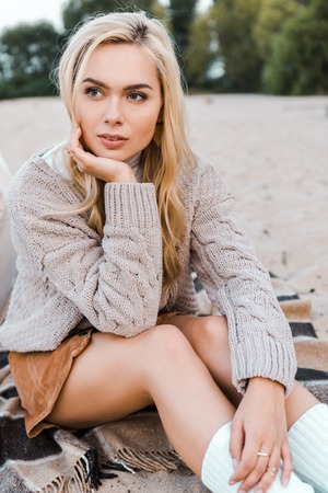 Attractive pensive blonde girl in autumn outfit sitting on beach and looking awayの写真素材