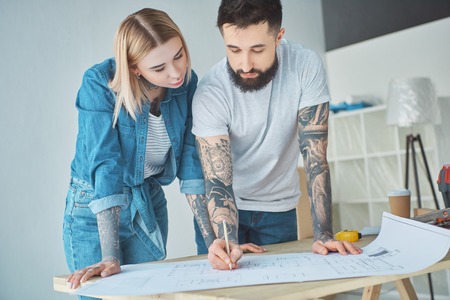 Portrait of tattooed couple looking at blueprints on wooden tabletop at new homeの写真素材