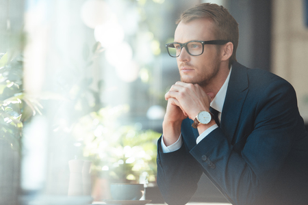 Thoughtful businessman in eyeglasses looking away in coffee shopの写真素材
