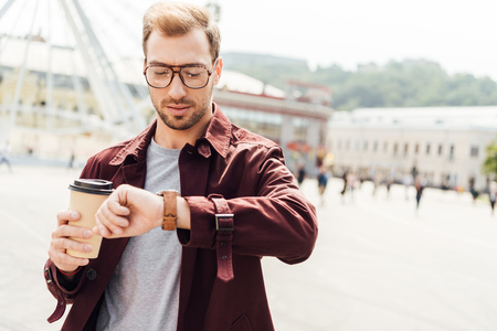 Handsome man in autumn outfit holding coffee in paper cup and checking time in cityの写真素材