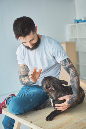 Tattooed man playing with french bulldog on wooden surface in new apartmentの写真素材