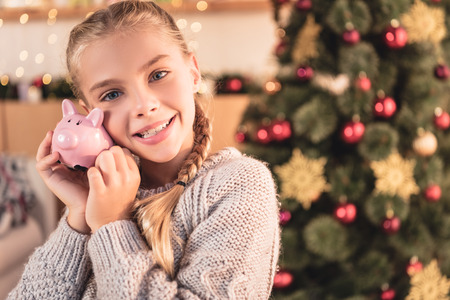 Happy kid holding piggy bank at home with Christmas treeの写真素材