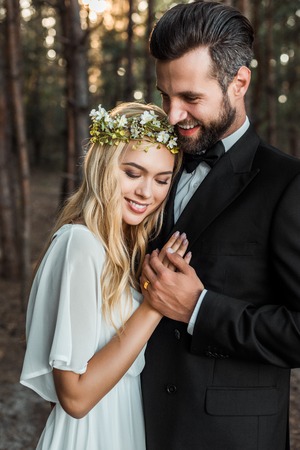 Beautiful happy bride in white dress and handsome groom in suit hugging and holding hands in forestの写真素材