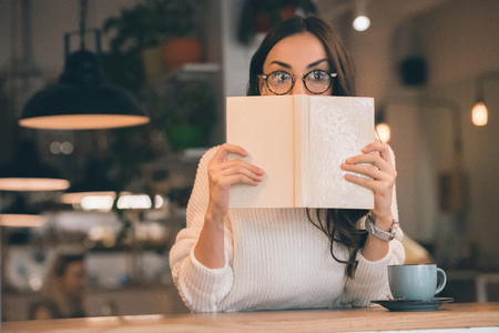 Shocked woman in eyeglasses covering face by book at table with coffee cup in cafe