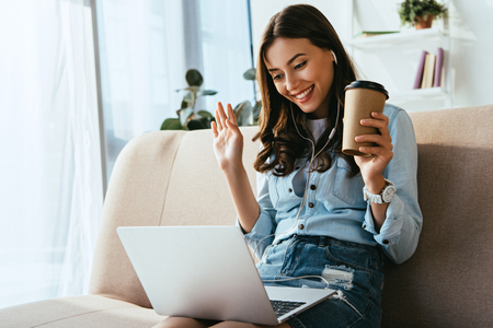 Smiling woman in earphones with coffee to go on sofa taking part in webinar at homeの写真素材