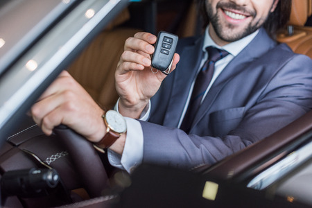 Partial view of smiling businessman with car key in hand sitting in new car in dealership salonの写真素材