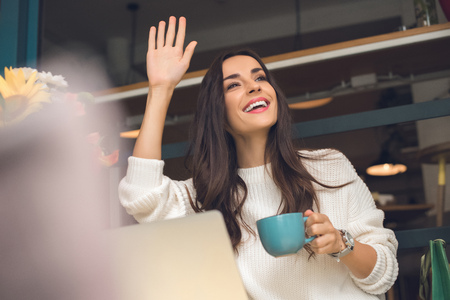 Happy female freelancer with coffee cup waving by hand at table with laptop in cafeの写真素材