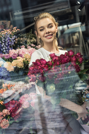View through window of attractive florist holding bouquet of burgundy roses in flower shop and looking at cameraの写真素材