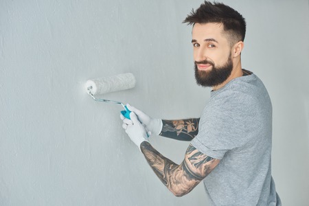 Smiling tattooed repairman with roll paint brush looking at camera while painting wall in new...