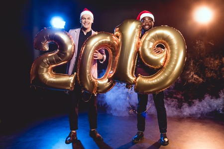 Happy multicultural friends in Santa hats holding 2019 new year golden balloons on smoke and backlitの写真素材