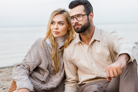 Stylish couple in autumn outfit sitting on beach and looking awayの写真素材