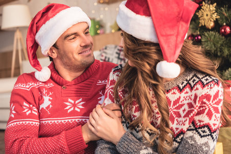Husband and wife in Santa hats holding hands and looking at each other at homeの写真素材