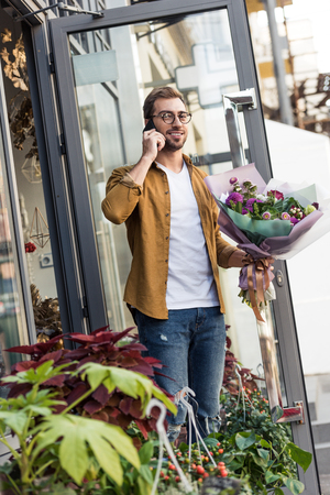 Smiling handsome man going out from flower shop with bouquet and talking by smartphoneの写真素材