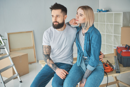 Young couple with tattoos sitting together and looking away in new homeの写真素材