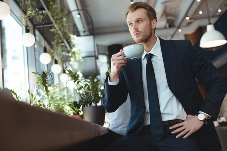 Stylish thoughtful businessman in suit with cup of coffee having coffee break in cafeの写真素材