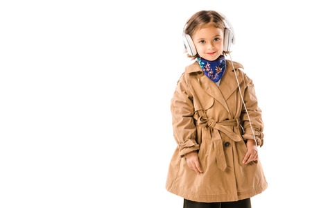 Smiling little child in stylish trench coat listening music with headphones and looking at camera isolated on white backgroundの写真素材
