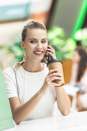 Beautiful smiling girl holding disposable coffee cup and talking by smartphone while sitting in cafe at shopping mallの写真素材