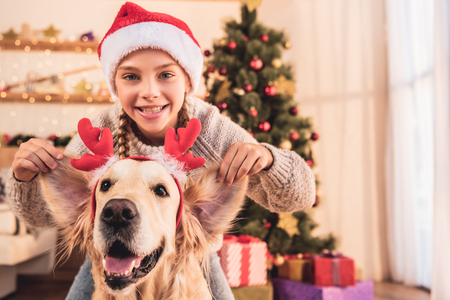 Smiling kid in Santa hat and golden retriever dog with deer horns having fun at home near Christmas treeの写真素材