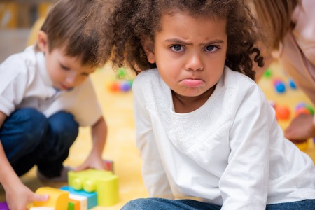 Dissatisfied African American kid grimacing and looking at camera in kindergarten