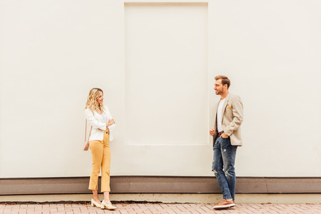 Couple in autumn outfit standing near beige wall on street and looking at each other