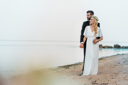 Handsome groom in suit hugging attractive bride in white dress on beachの写真素材