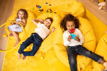 High angle view of multiethnic kids lying on bean bag chairs with toys in kindergartenの写真素材